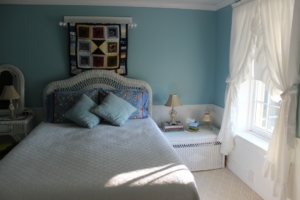 The cottage room with a white bedspread and light blue walls. A quilt hanging on the wall
